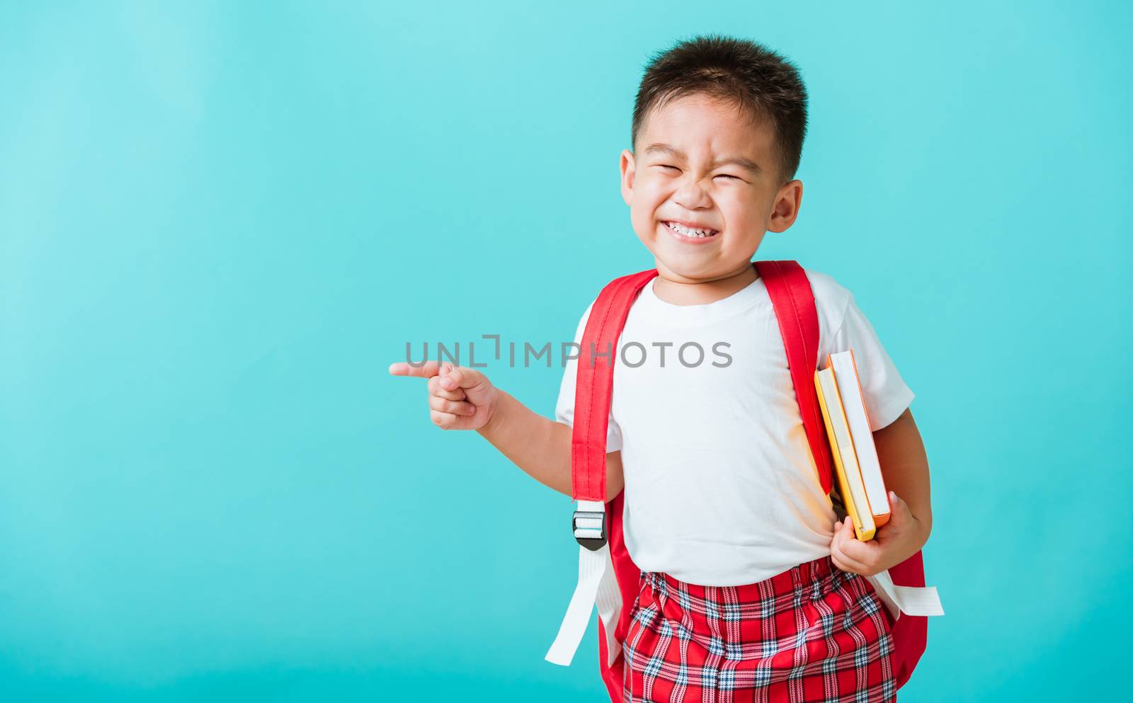 Back to school concept. Portrait Asian happy funny cute little child boy smile hug books and point finger to side away space, isolated blue background. Kid from preschool kindergarten with school bag