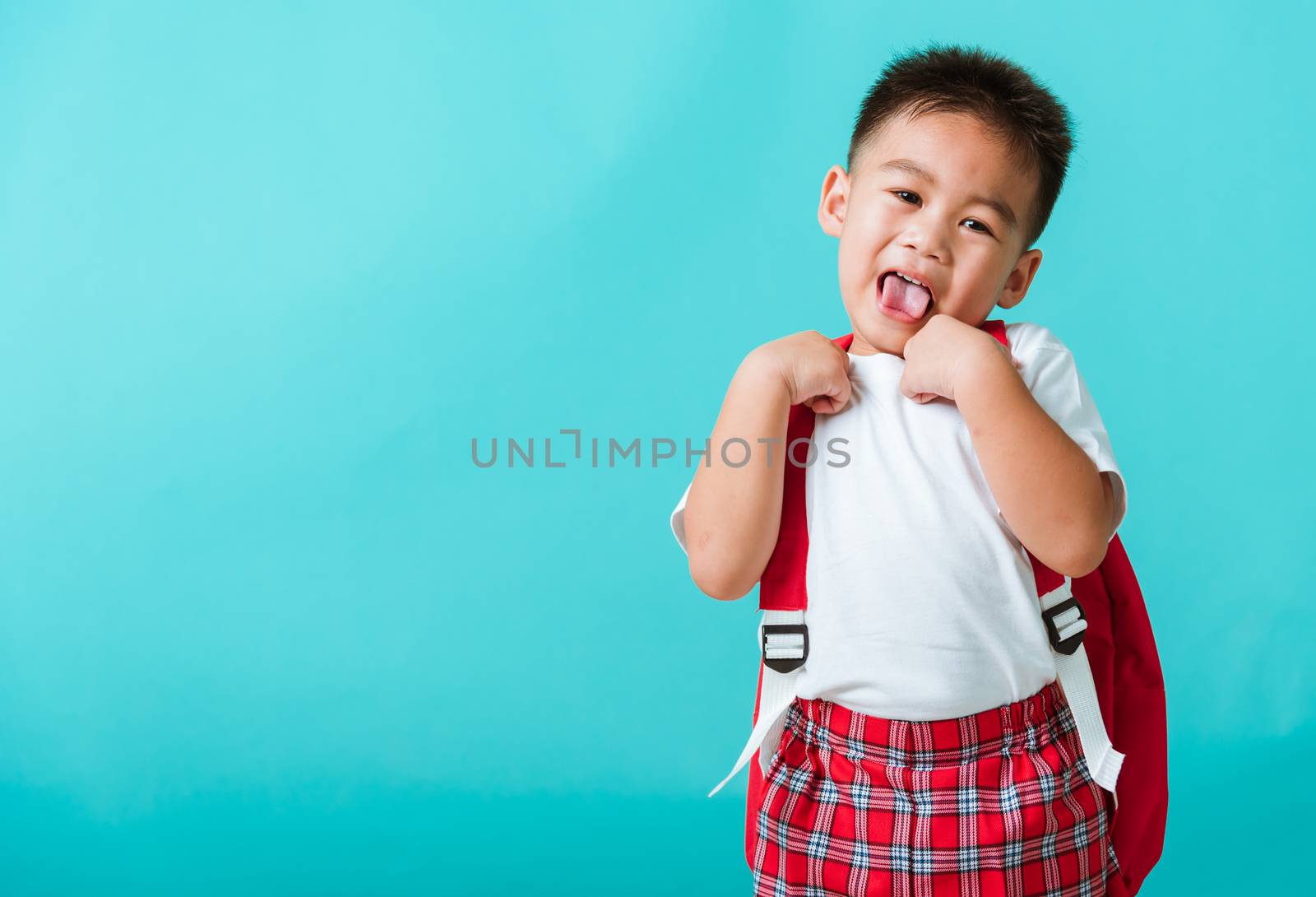 Back to school concept. Portrait closeup happy Asian cute little child boy in uniform smiling, isolated blue background. The kid from preschool kindergarten with a school bag backpack