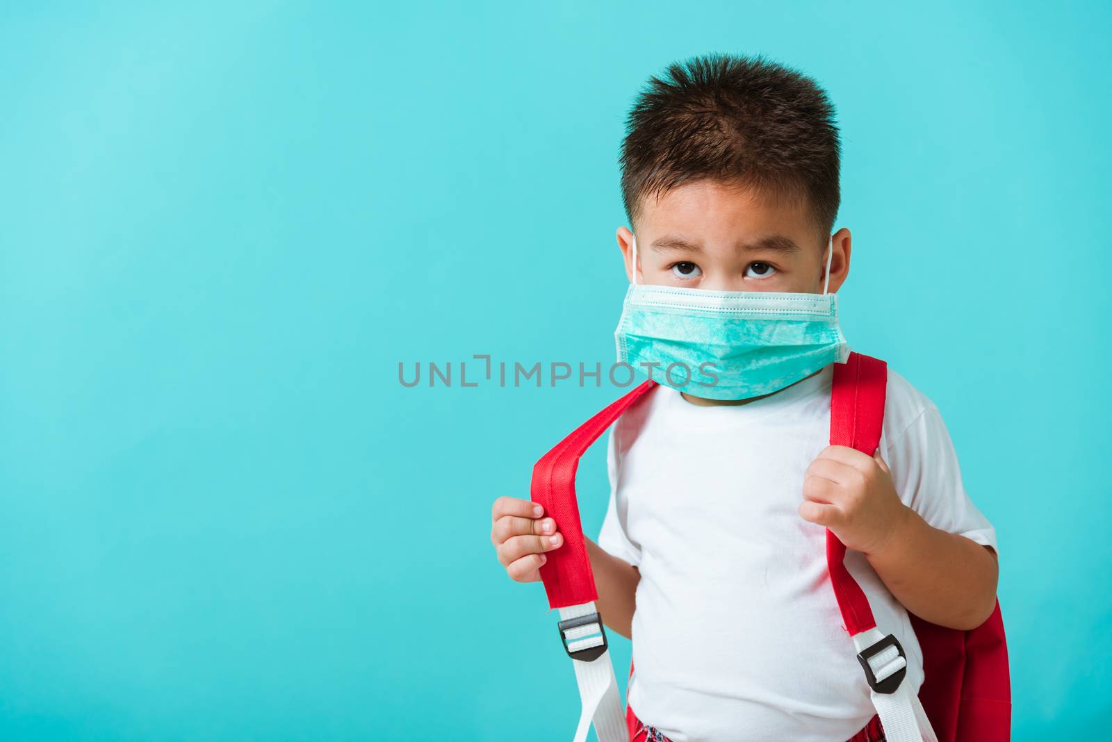 Back to school coronavirus Covid-19 education. Portrait Asian cute little child boy kindergarten wear face mask protective and school bag before going to school, studio shot isolated blue background