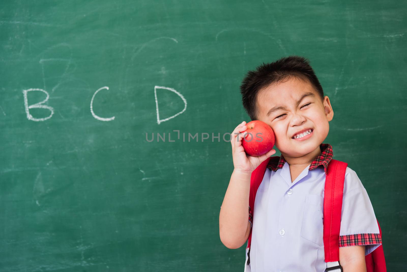 Back to School. Happy Asian funny cute little child boy from kindergarten in student uniform with school bag hold red apple on hand smiling on green school blackboard, First time to school education