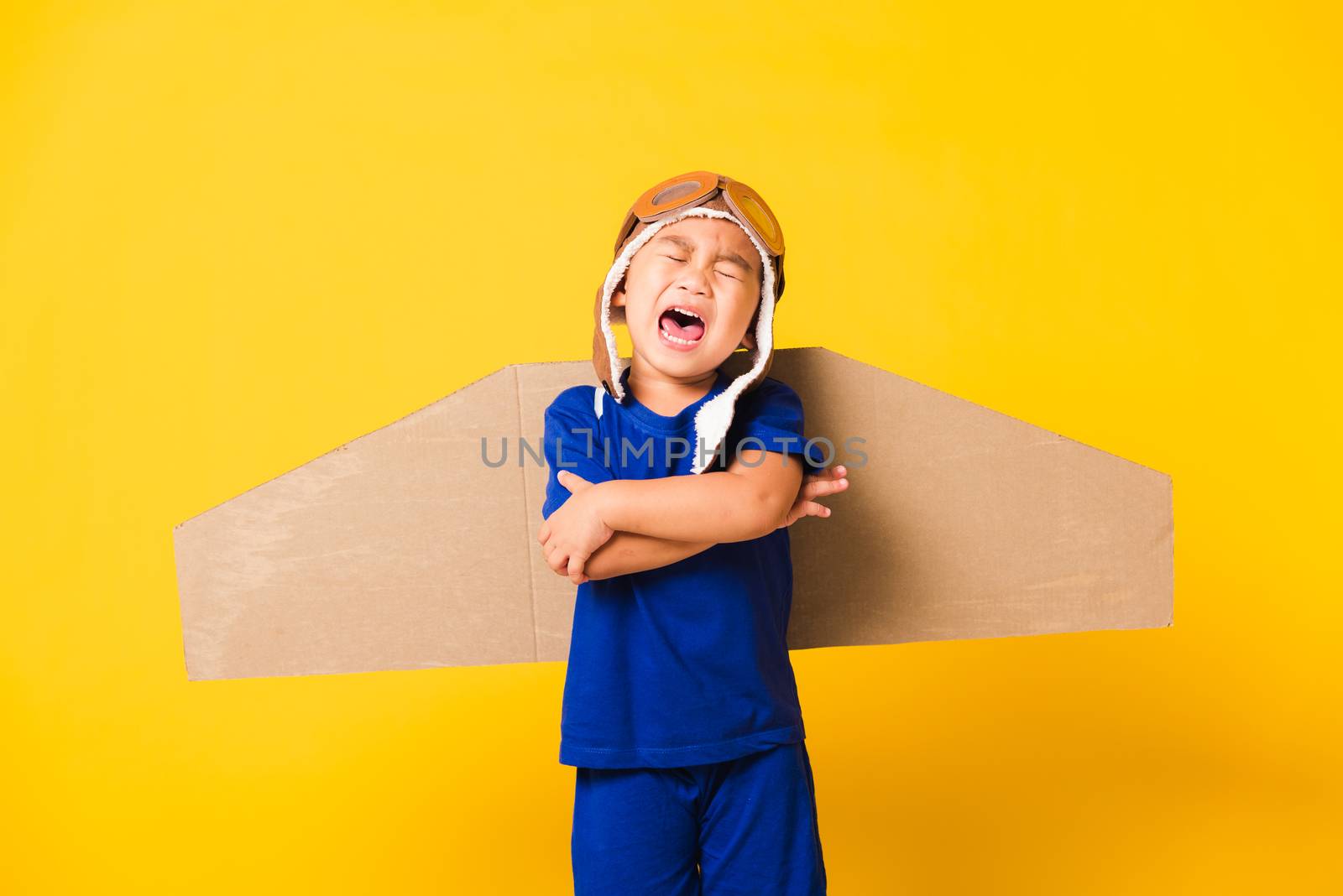 Happy Asian handsome funny child or kid little boy smile wear pilot hat play and goggles with toy cardboard airplane wings flying crossed arm, studio shot isolated yellow background, Startup freedom