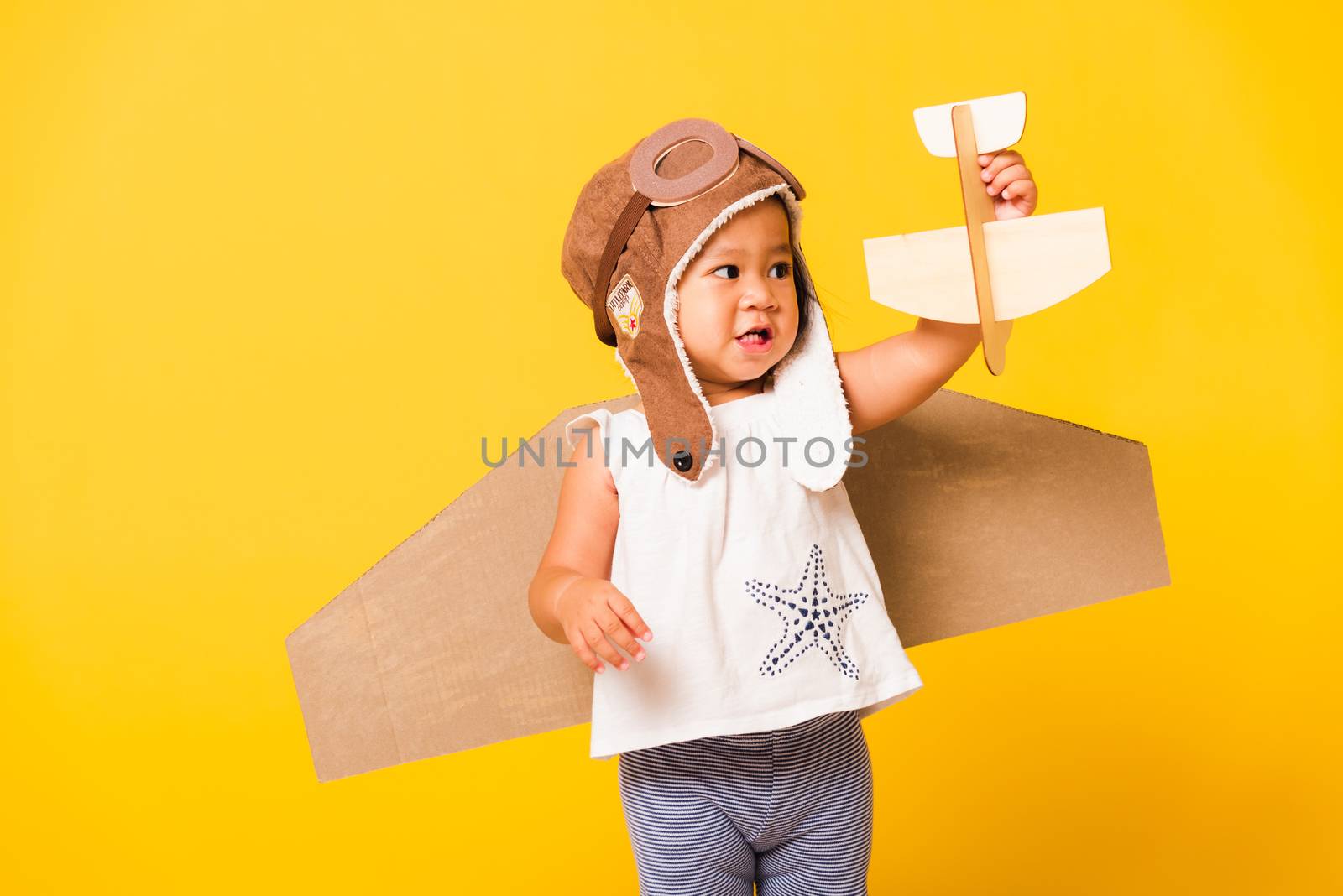 Happy Asian beautiful funny baby little girl smile wear pilot hat play and goggles with toy cardboard airplane wings fly hold plane toy, studio shot isolated yellow background, Startup freedom concept