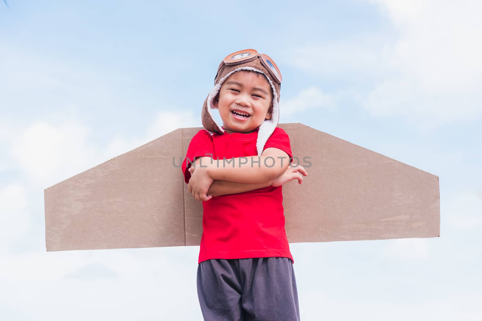 Happy Asian funny child or kid little boy smile wear pilot hat and goggles play toy cardboard airplane wing flying stand crossed arm against summer blue sky cloud background, Startup freedom concept