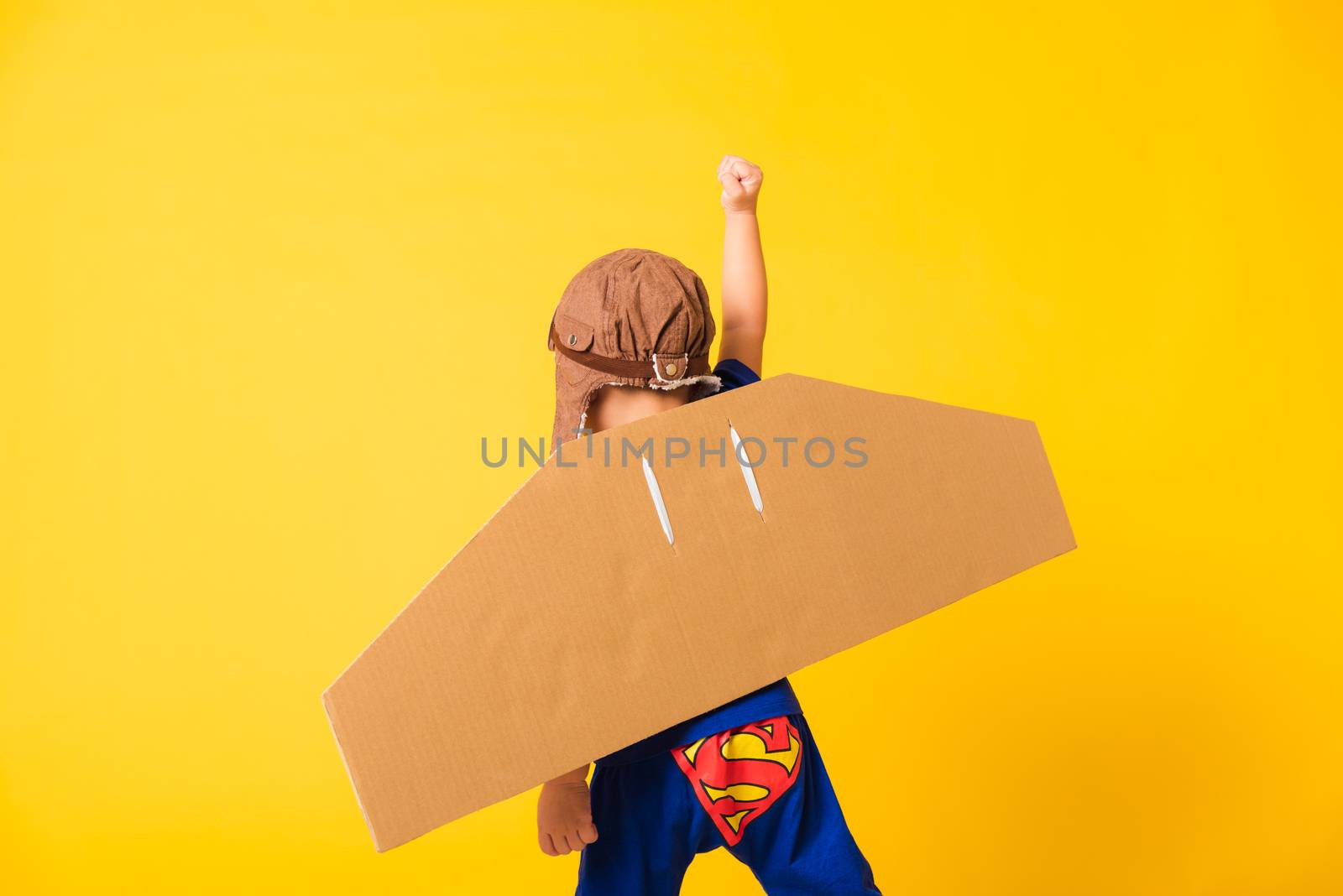 Back of Happy Asian handsome funny child or kid little boy smile wear pilot hat play raise hand up with toy cardboard airplane wings flying, studio shot isolated yellow background, Startup freedom