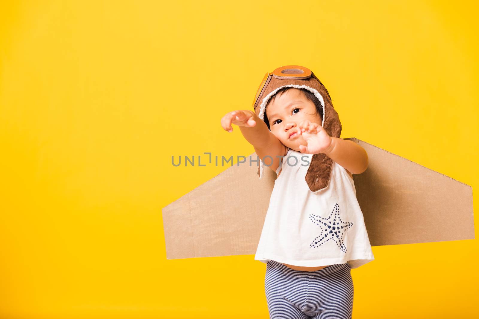 Happy Asian beautiful funny baby little girl smile wear pilot hat raise hand up play and goggles with toy cardboard airplane wings fly, studio shot isolated yellow background, Startup freedom concept