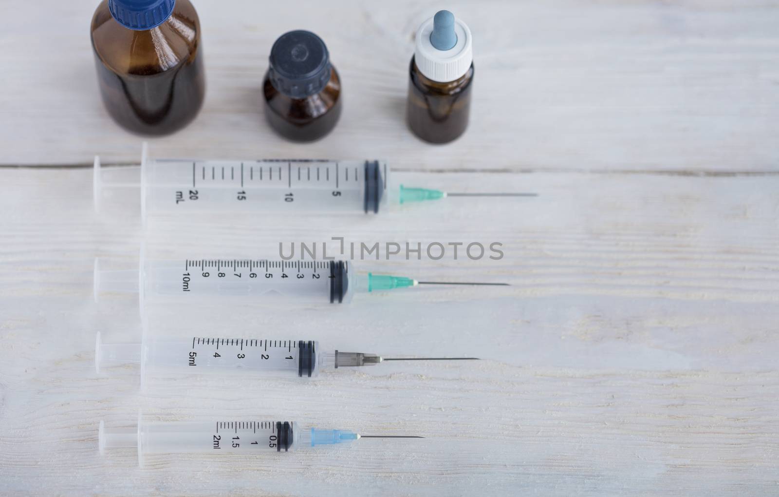 Syringes, capsules and medical bottles lie horizontally on a white roughly painted wooden table.