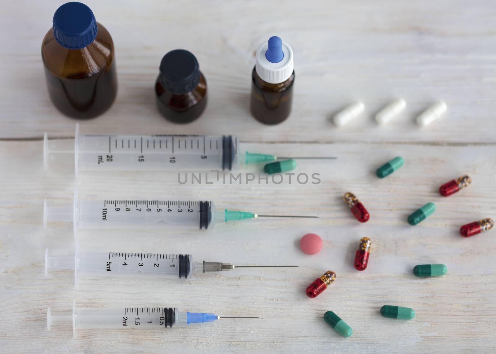 Syringes, capsules, pills and medical bottles lie horizontally on a white roughly painted wooden table.