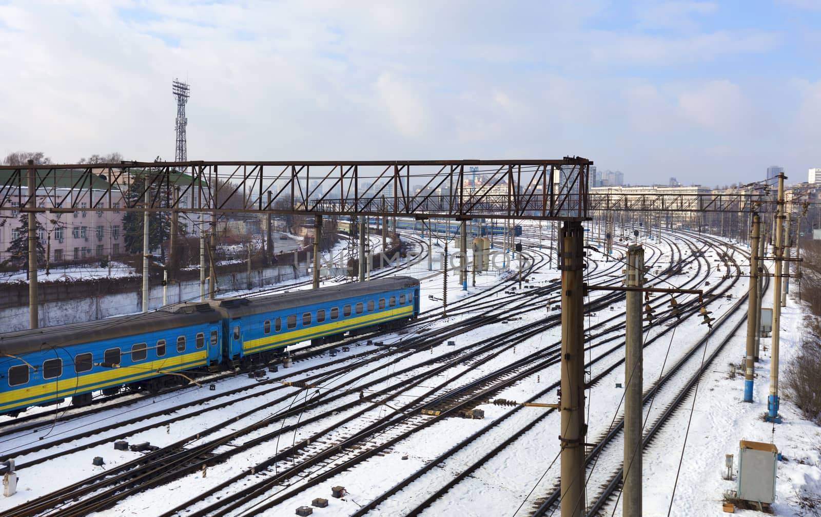 Passenger railway wagons ride along the railway tracks in the winter season against the backdrop of the cityscape by Sergii