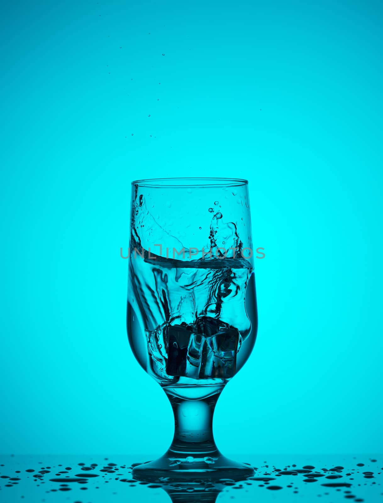 Splash of water from a falling ice cube into a transparent glass glass on a turquoise background color
