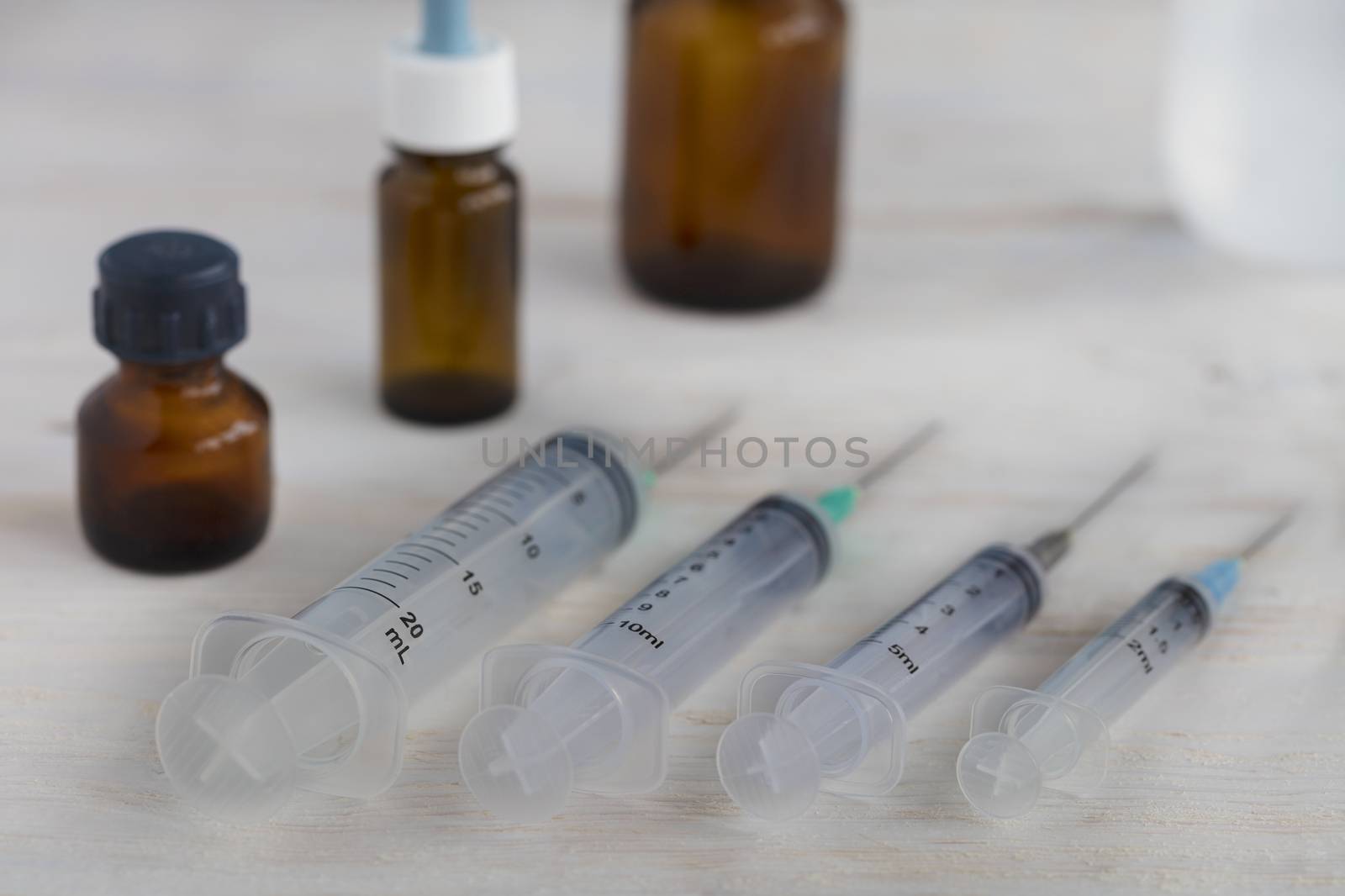 Medical syringes of various capacities and medical bottles lie diagonally on a white roughly painted wooden table.