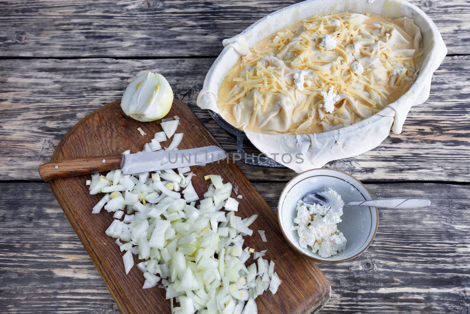 Ingredients for vegetable pie: chopped onions with knife, dough, feta, cheese on an old wooden weathered table.