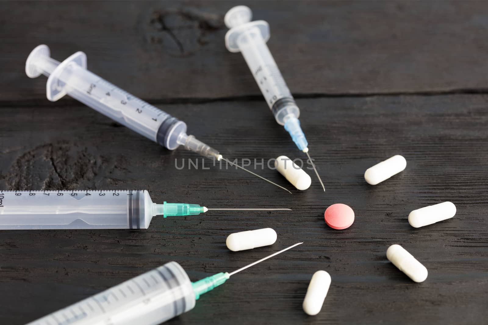Medical syringes of various capacities, capsules, tablets lie on an old black wooden board and point to a red round tablet.