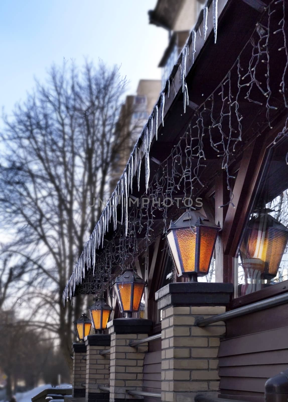 Four old street lamps shine near the restaurant windows in the open air in the early spring morning. Icy icicles look down on them.