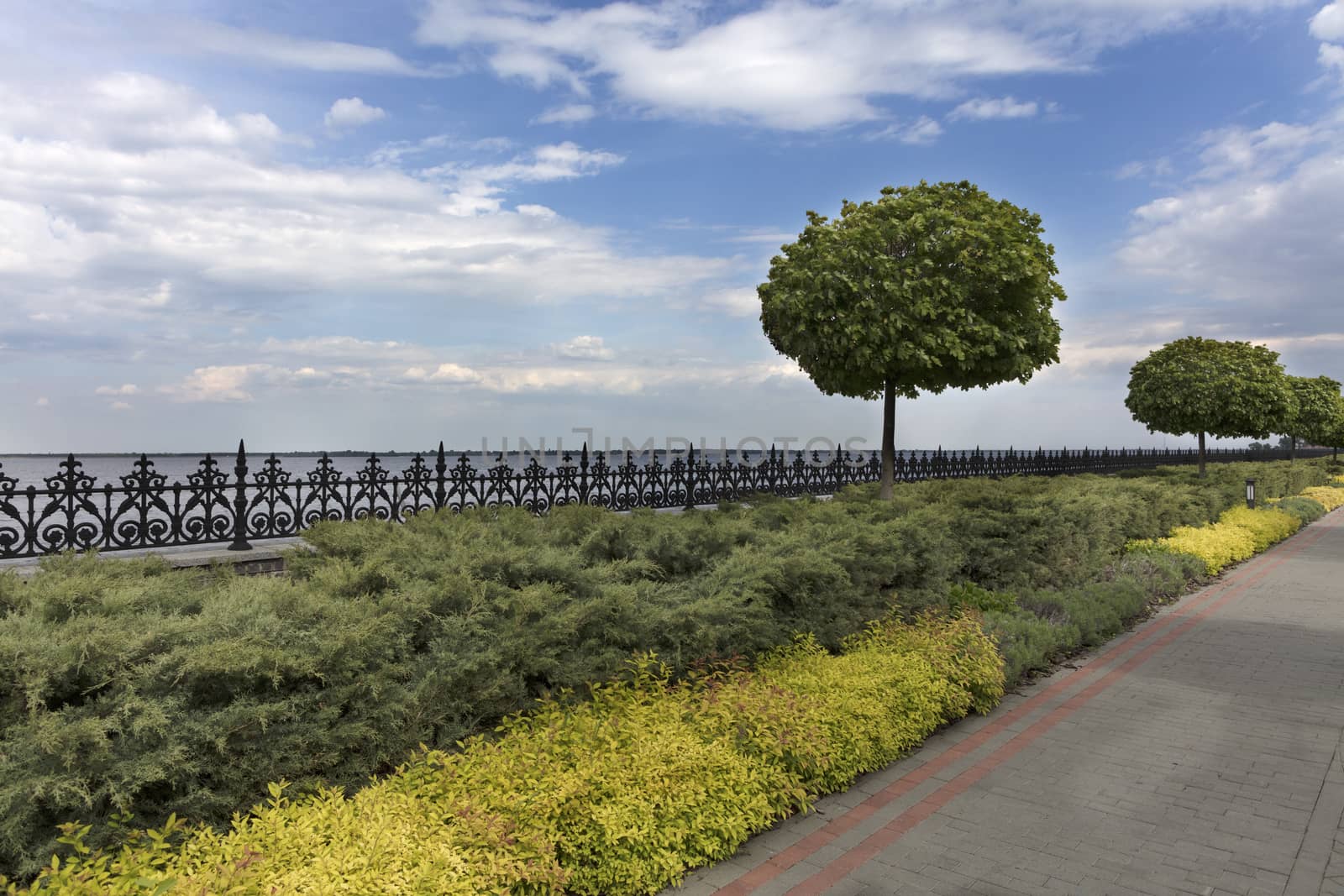 A paved stepped road in a beautiful park passes along the embankment of the river with ornamental flowers, framed decorative fence with cut bushes and lanterns for lighting.