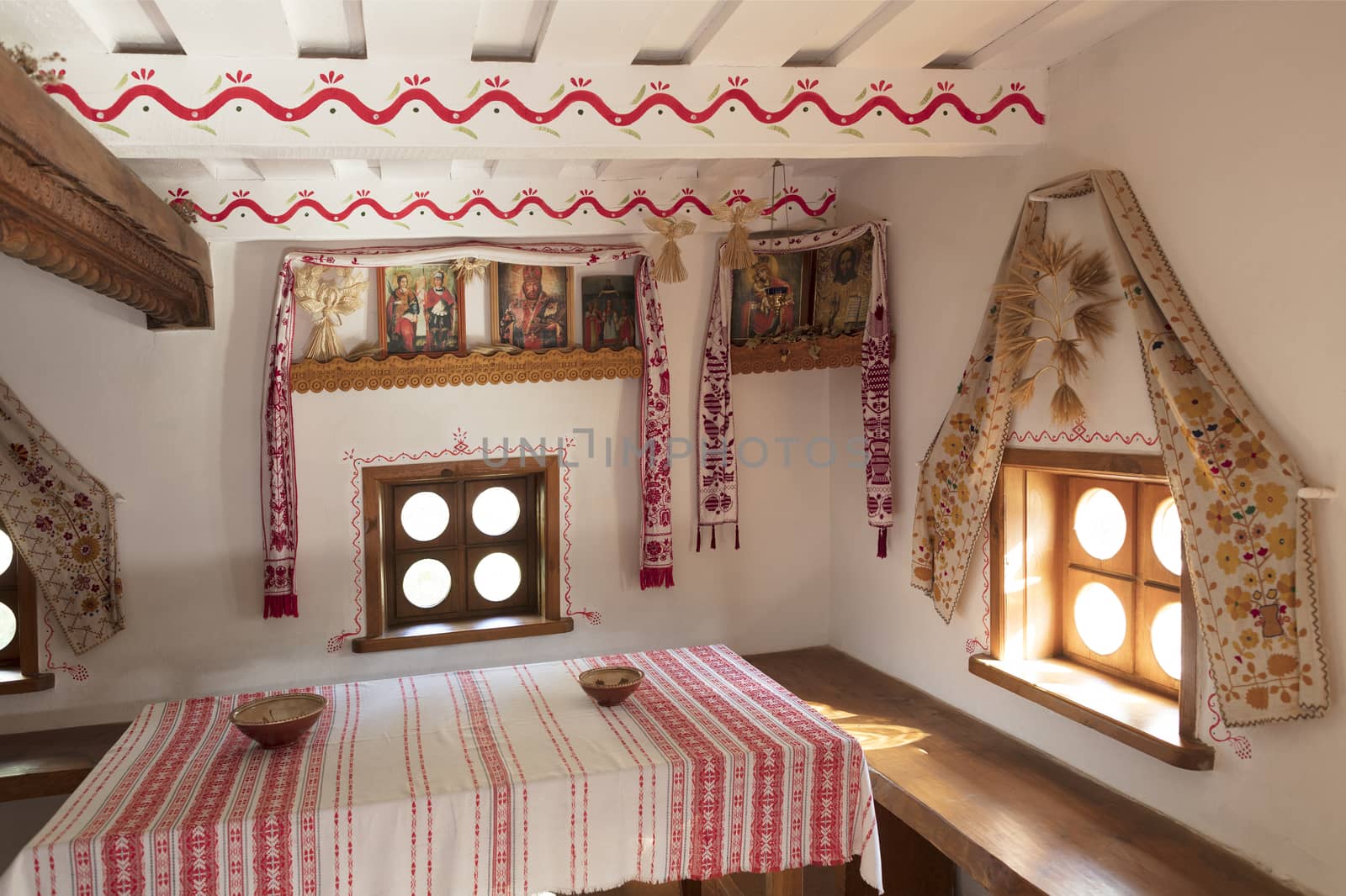 Ceramic bowls, plates and embroidered tablecloth on a wooden table, wooden lavas around the perimeter of the room, embroidered towels and icons on the walls in the interior of the ancient Ukrainian rural house. Ukraine, Kyiv, 18 century.