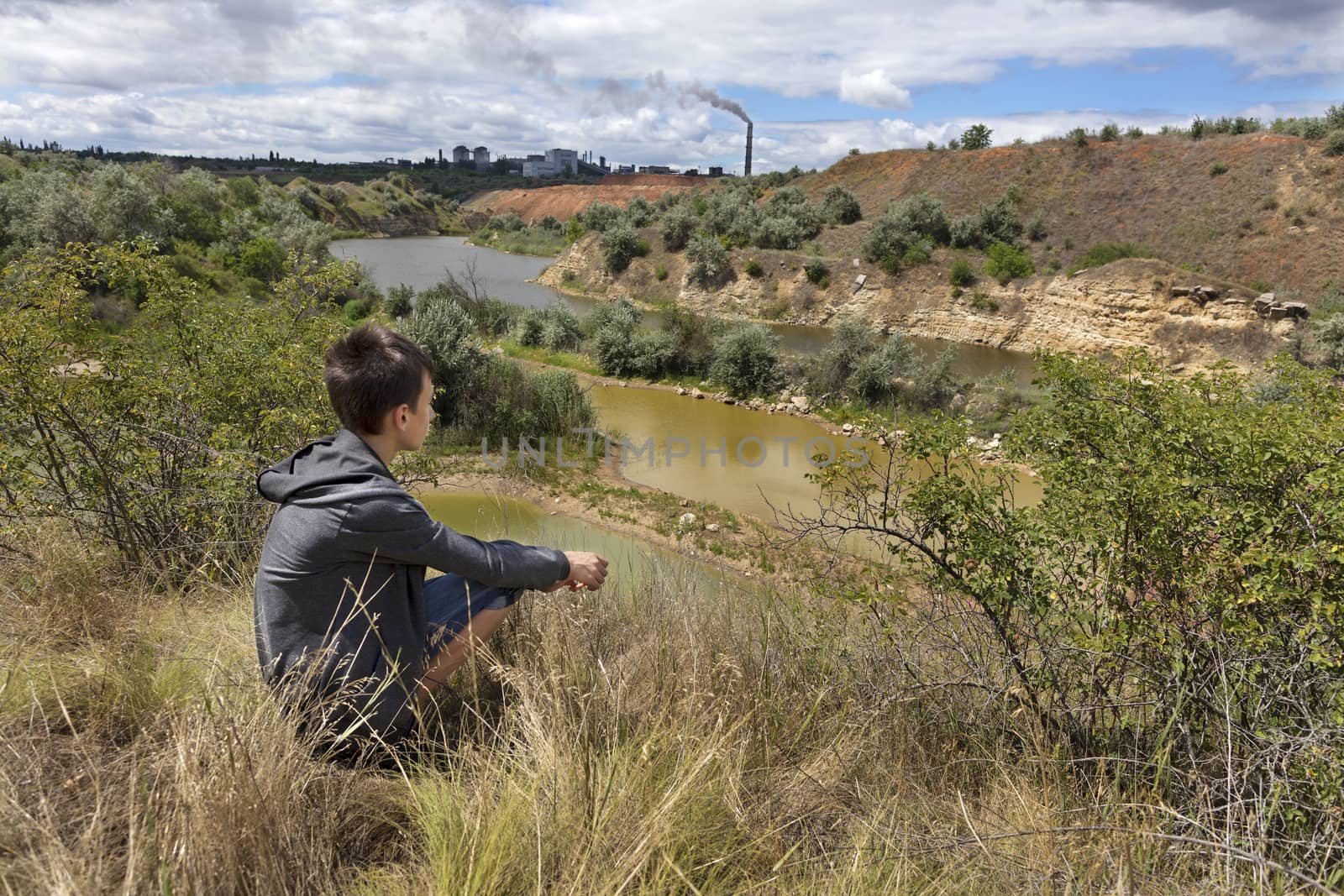 A teenager looks at the landscape of an abandoned sandy quarry on a summer day. Recycling company on the horizon.