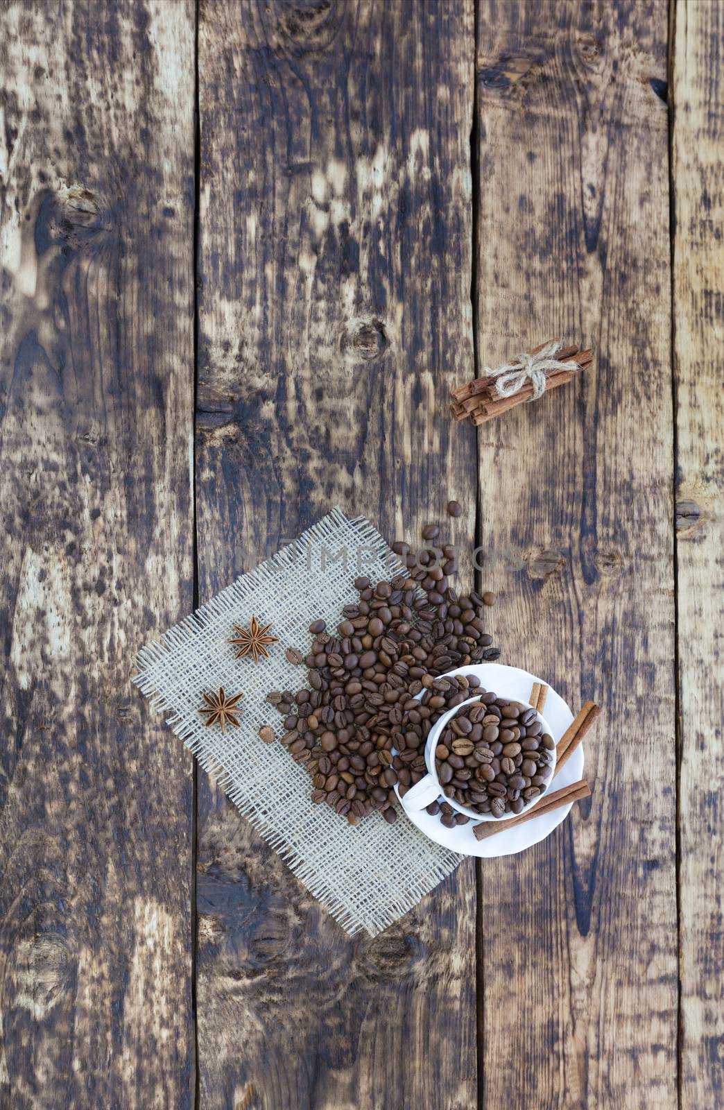 On the old wooden table is a white cup with grains of coffee. Grains of coffee are scattered on a linen napkin. The cinnamon is tied with a rope. Anise complements the aroma of coffee. Top view, vertical placement.
