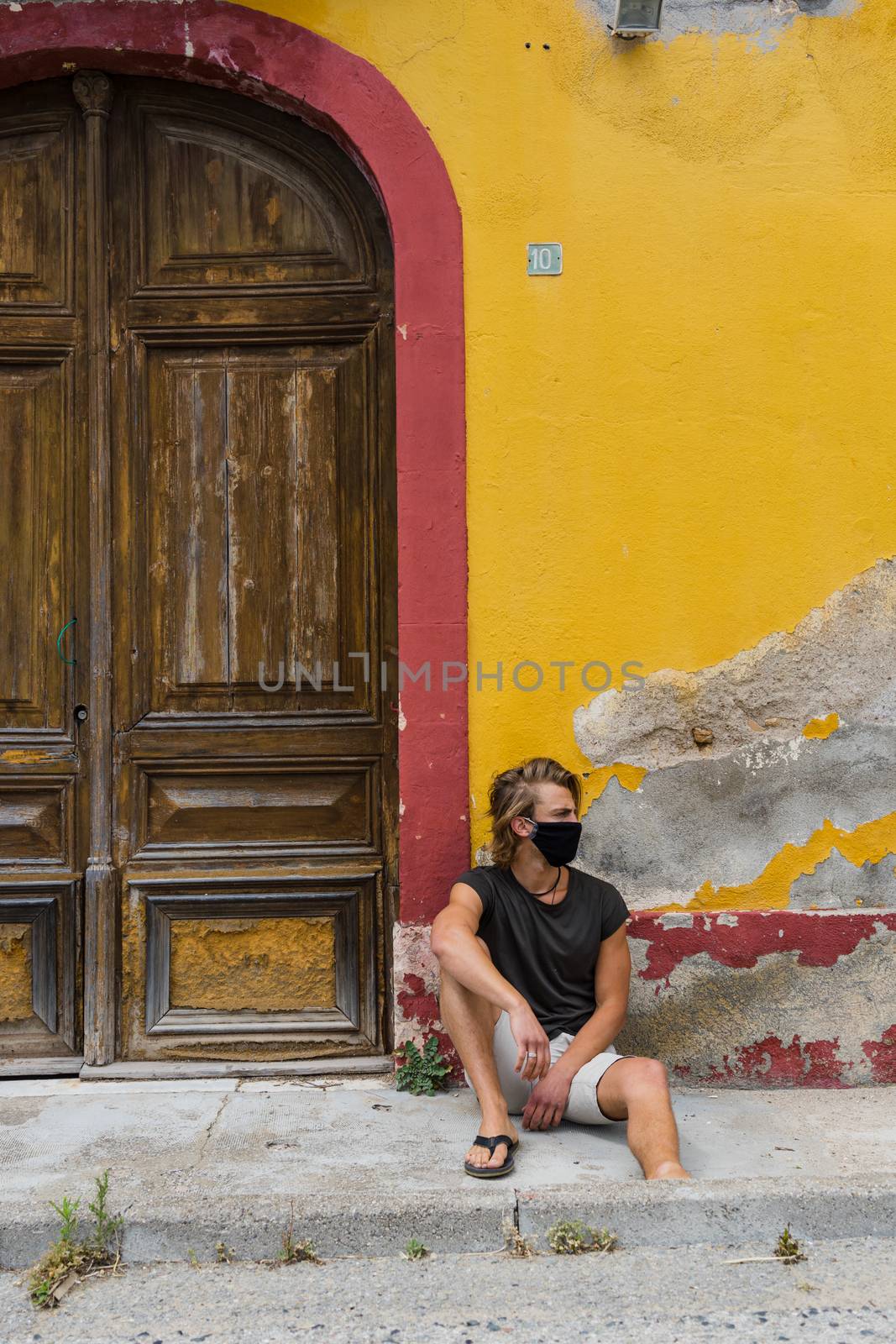 Young blond man with medium long hair with mask in his mouth from the virus, sitting on the street in front of a rustic old house