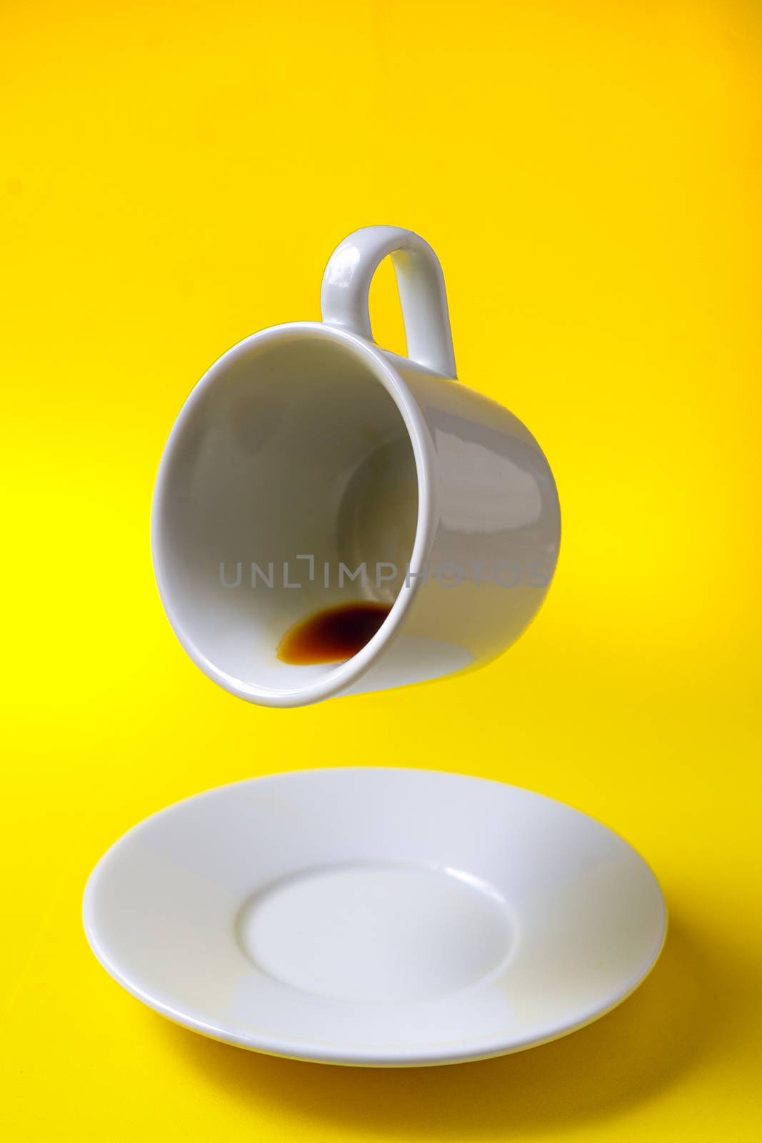 white cup of coffee with coffee floating tilted in the air and a saucer on a yellow background