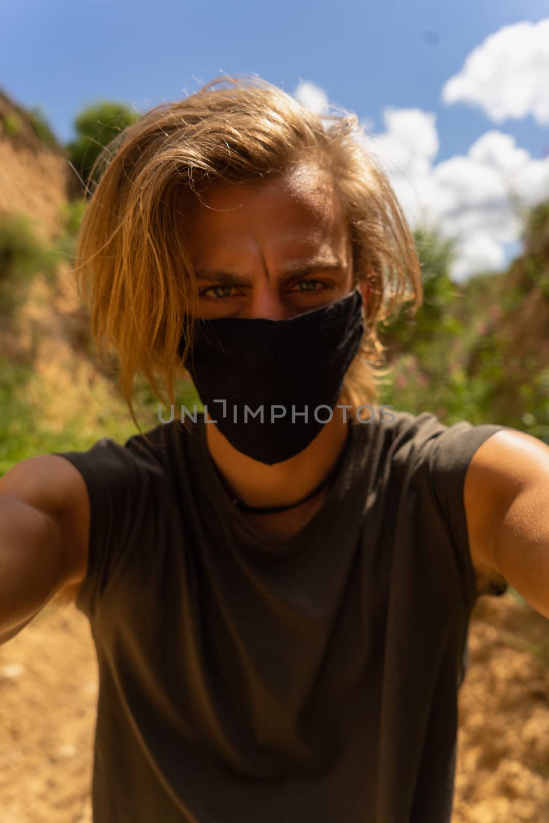 Self portrait of young man with medium long blond hair and blue eyes with mask in his mouth from the virus in the middle of the forest and nature