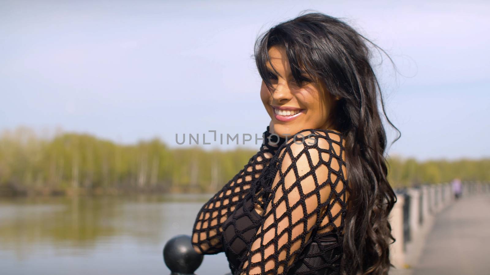 Close up woman with beautiful hair posing near the river. Portrait of smiling lady in black dress on the embankment in the city park. Real people