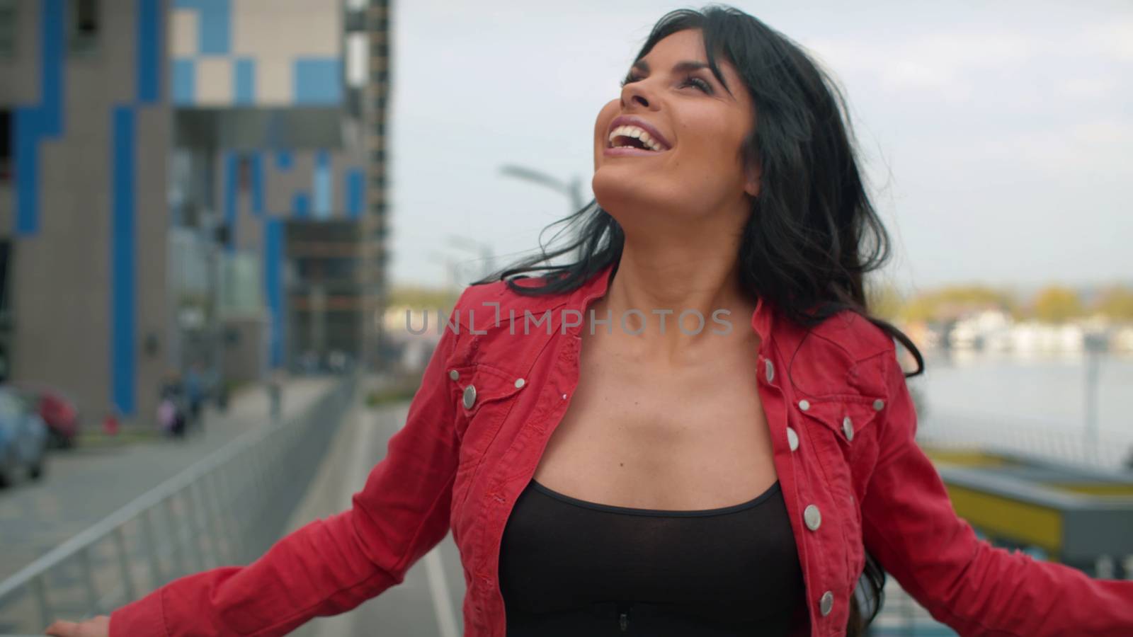 Close up portrait of laughing beautiful woman with long black hair standing next to a modern building. Girl in red jacket in the city. Real people