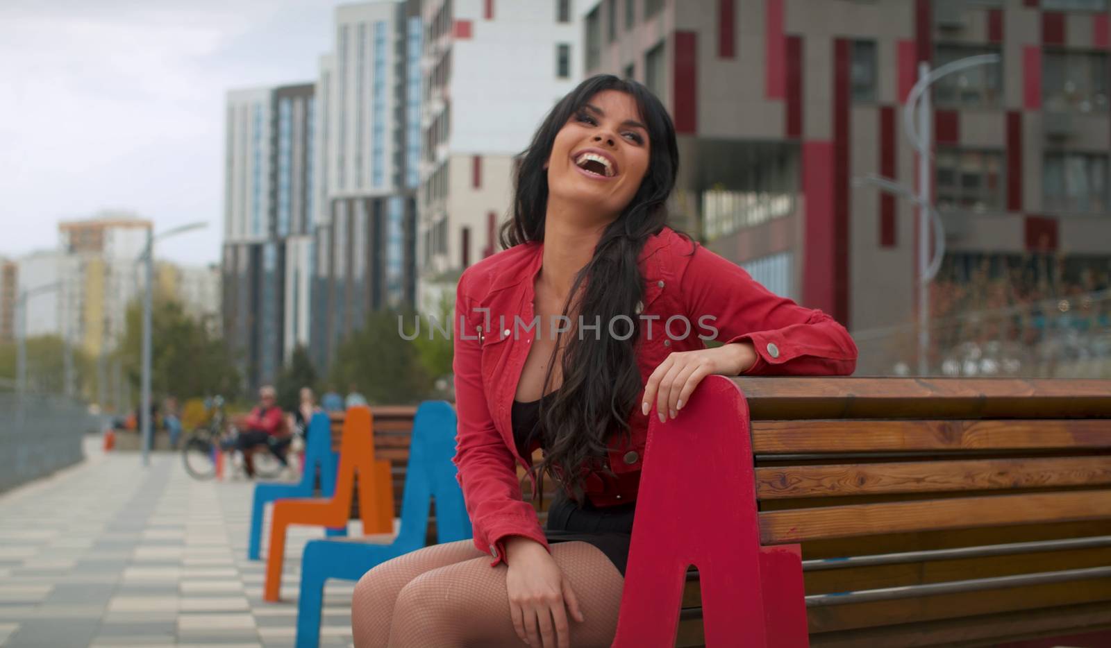 Close up portrait of beautiful brunette laughing woman in red jacket sitting on the bench outdoors. Real people in the city.