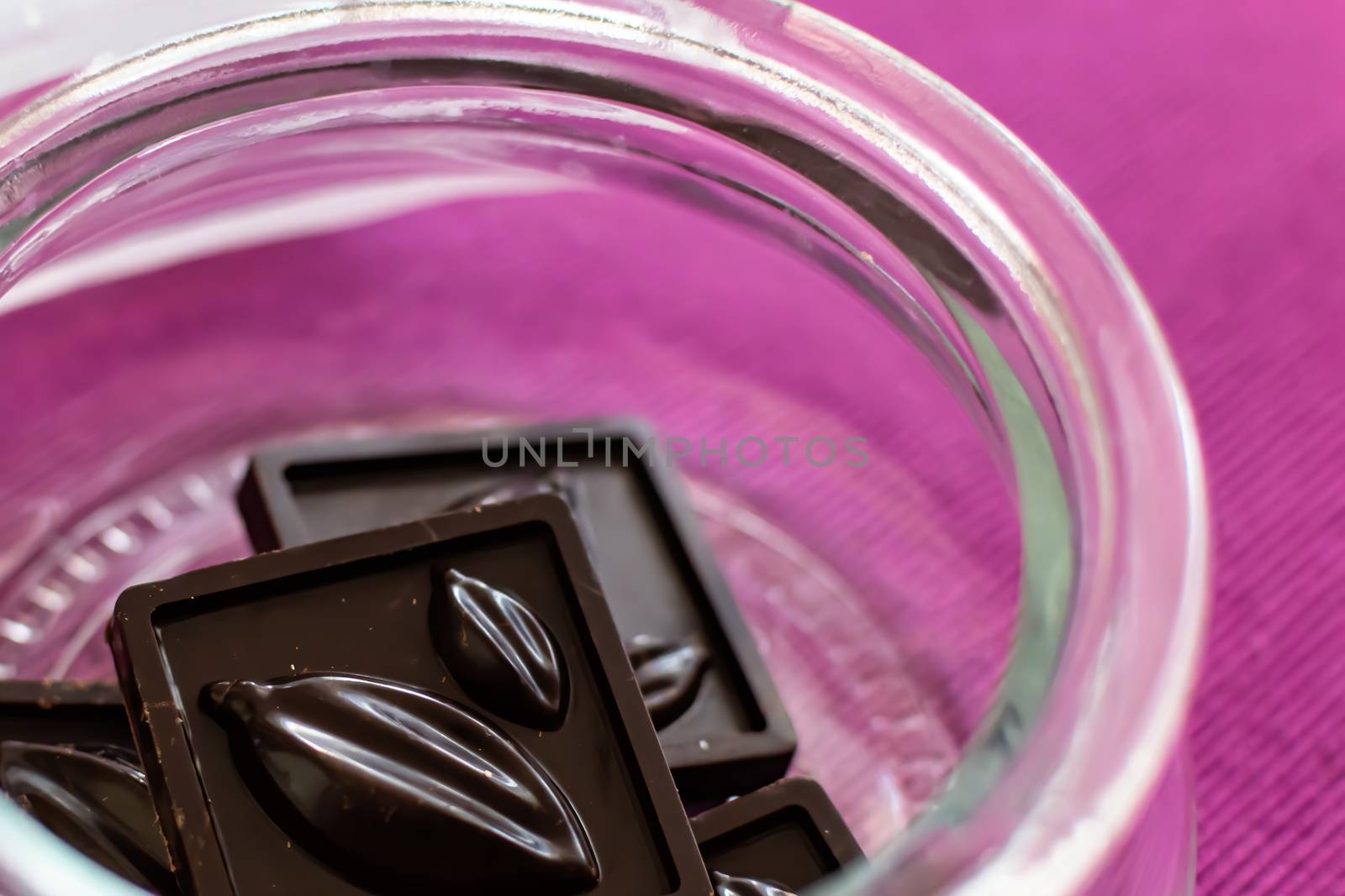 Pieces of handmade chocolate are in a glass jar on a lilac background, close-up, selective focus. Useful and delicious sweets.