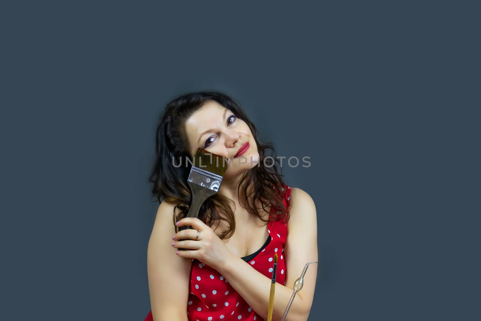 Portrait of a beautiful, fashionable middle-aged woman with long dark hair in a red dress, holding a paintbrush and posing on a dark gray background. Free space to copy your design.