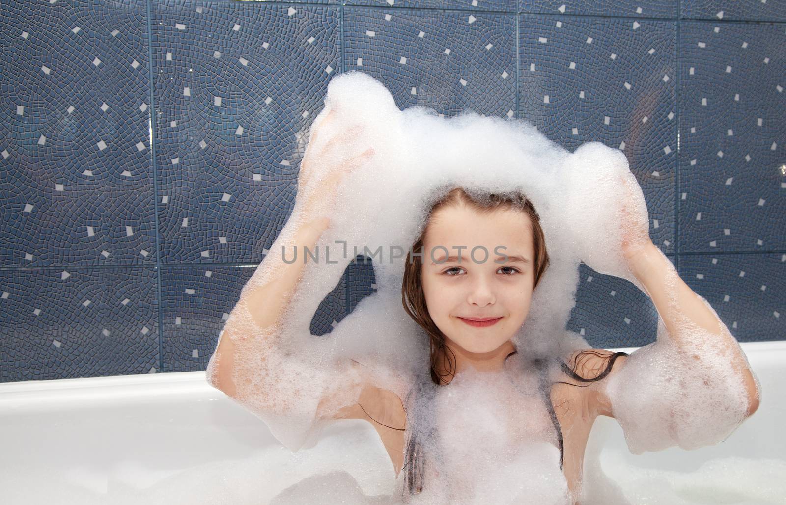 smiling little girl sitting in a bath and imposes soap suds on her head
