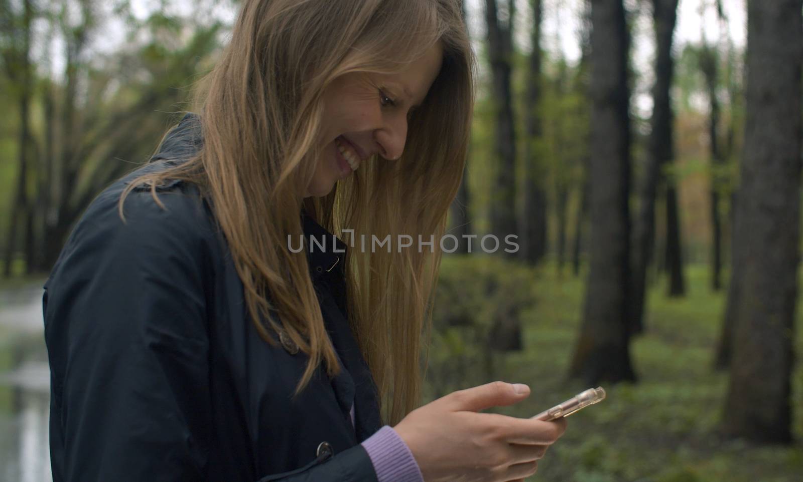 Attractive young woman reading something in smart phone and laughing. Blond cheerful woman on the stroll in the park in spring.