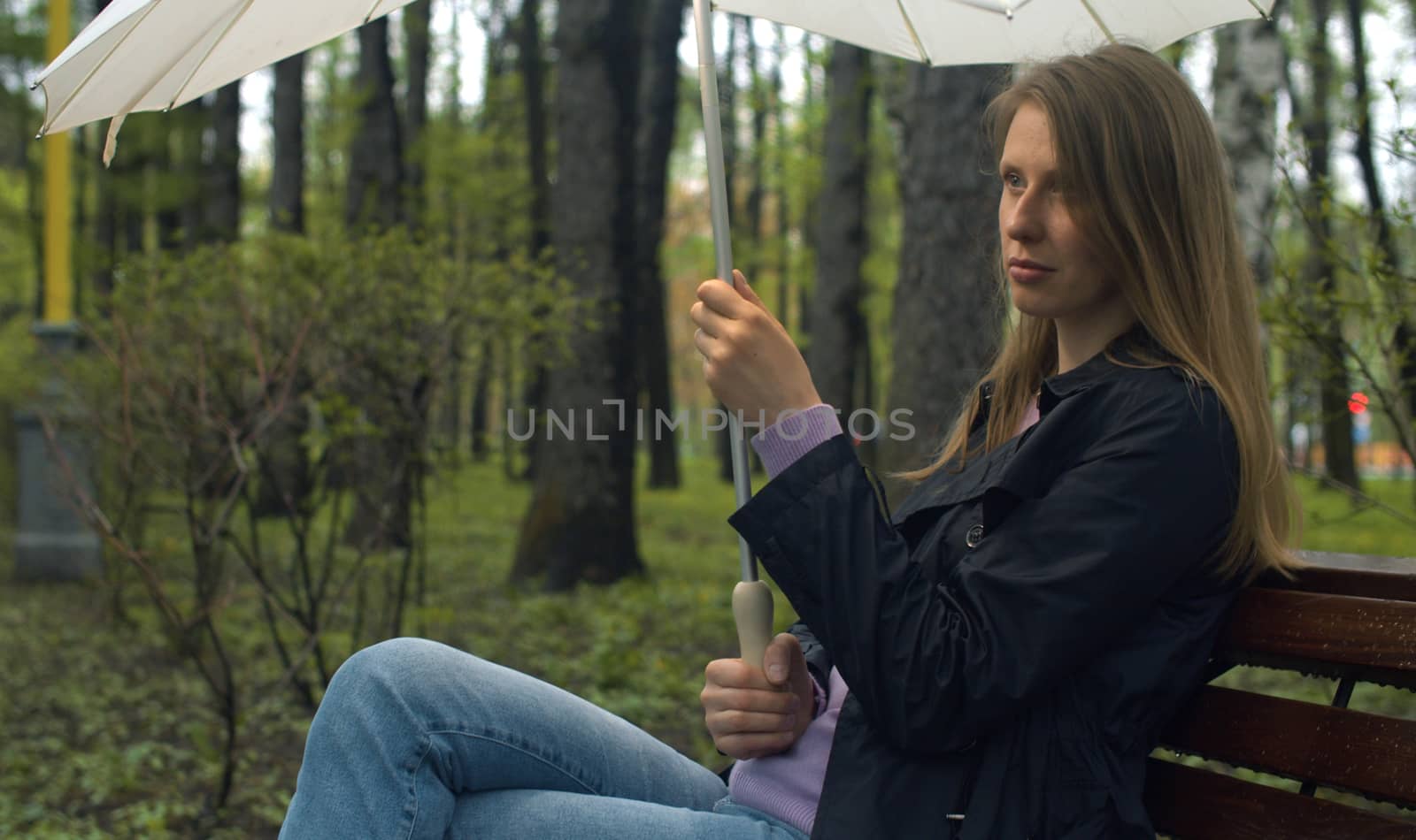 Portrait of a beautiful serious woman with umbrella sitting on the bench in the park in spring. Rainy weather, real people