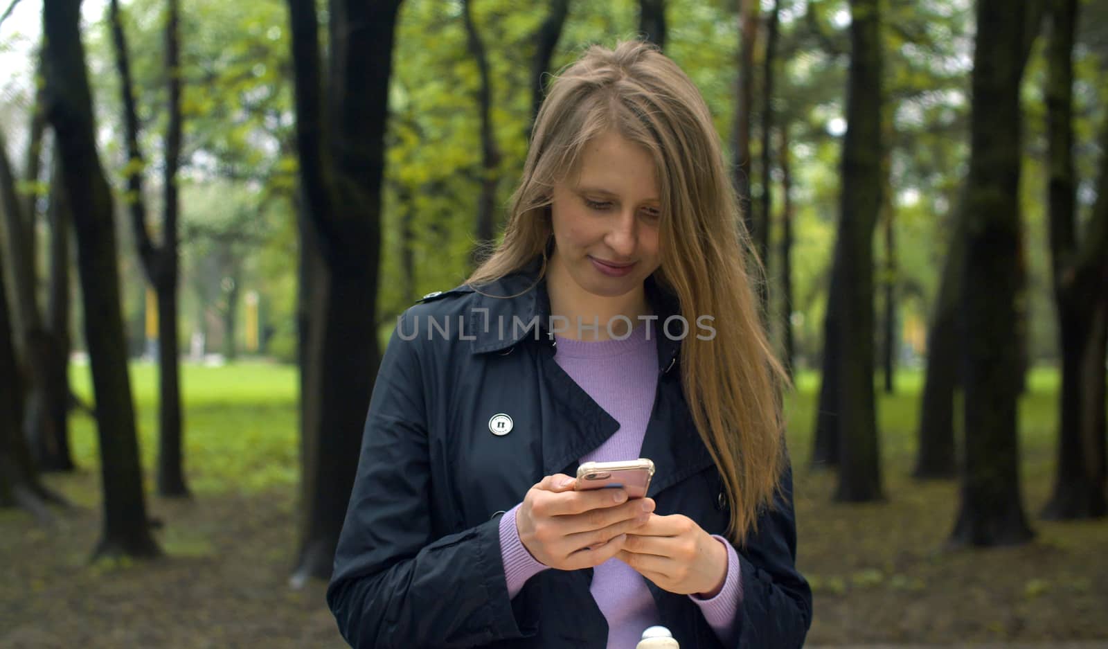 Attractive young woman reading something in smart phone and laughing. Blond cheerful woman on the stroll in the park in spring//