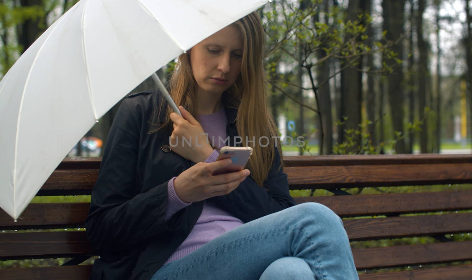 Portrait of a beautiful serious woman with umbrella sitting on the bench in the park in spring. Rainy weather, real people