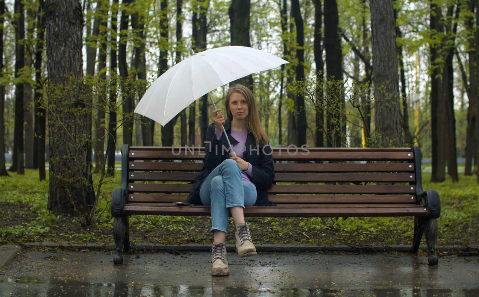 Portrait of a beautiful pensive woman with umbrella sitting on the bench in the park in spring. Rainy weather, real people