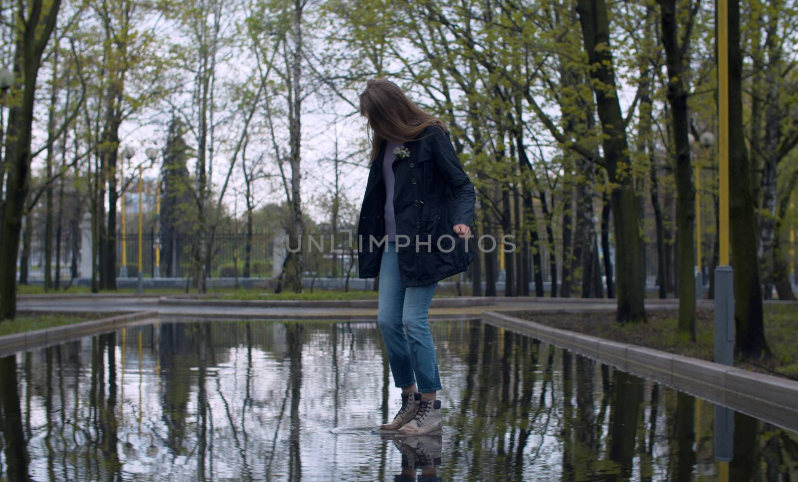 Happy woman jumping in a deep puddle in large splashes. Trees are reflected in a puddle. Rainy day in the park. Real people