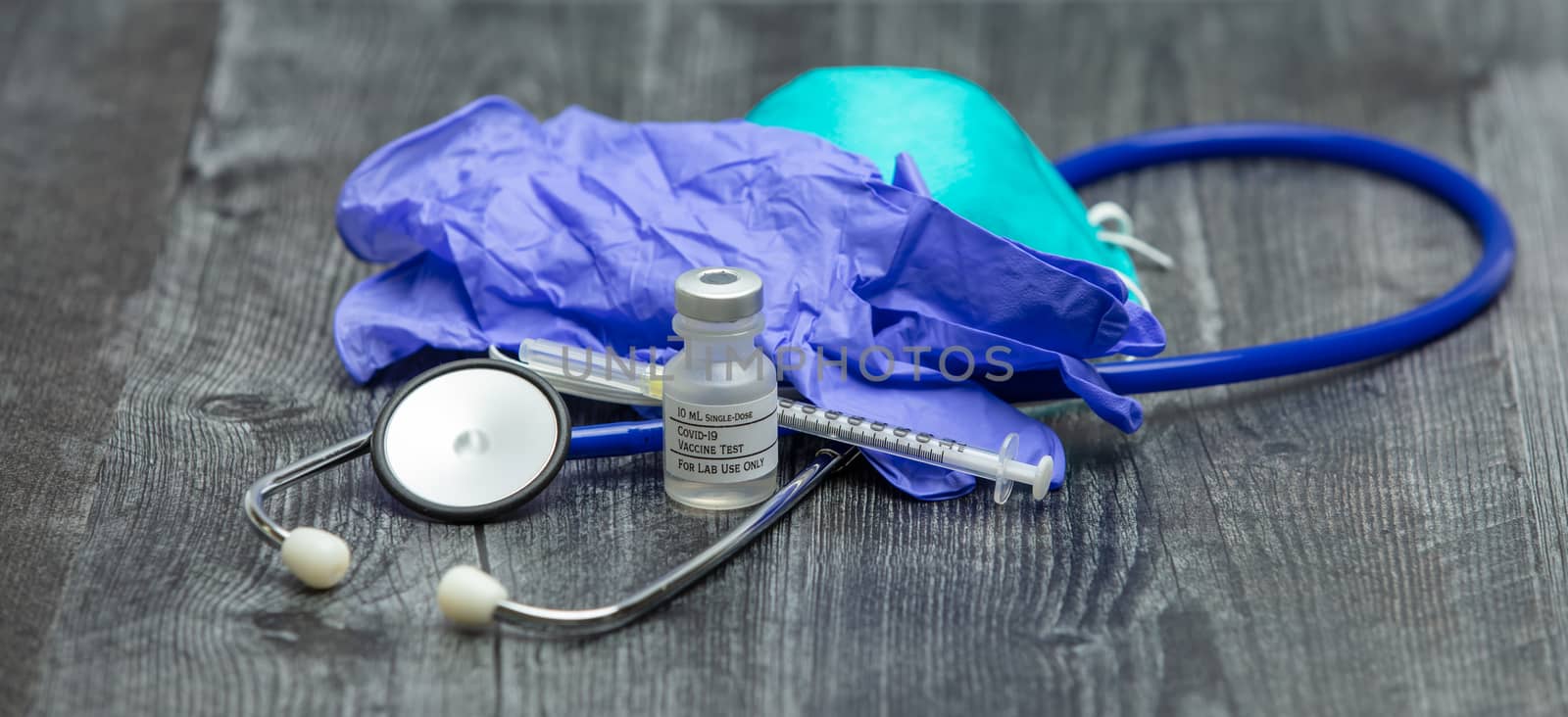A blue medical stethoscope, face mask respirator, gloves, syringe and vial of covid-19 test vaccine on a wooden surface.