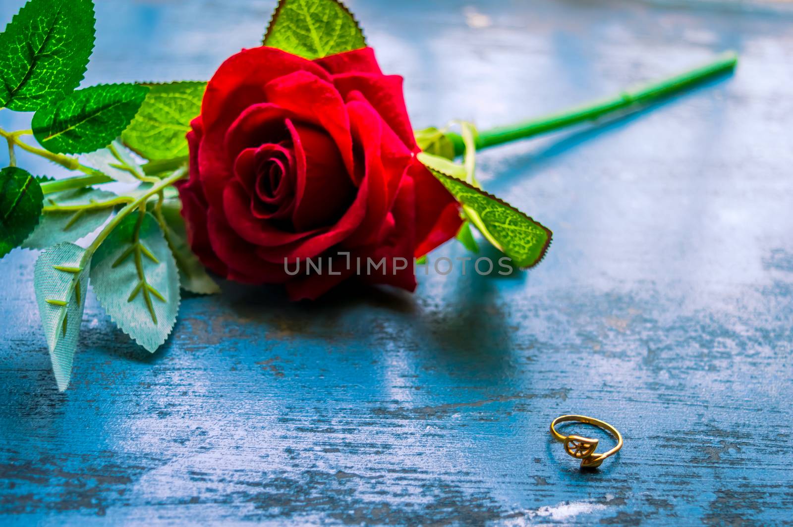 Close up gold engagement ring jewelry on rustic metal floor. Soft focus romantic Red rose flower in background. Love Proposal or Propose concept for valentines day wedding and holidays. Copy Space.