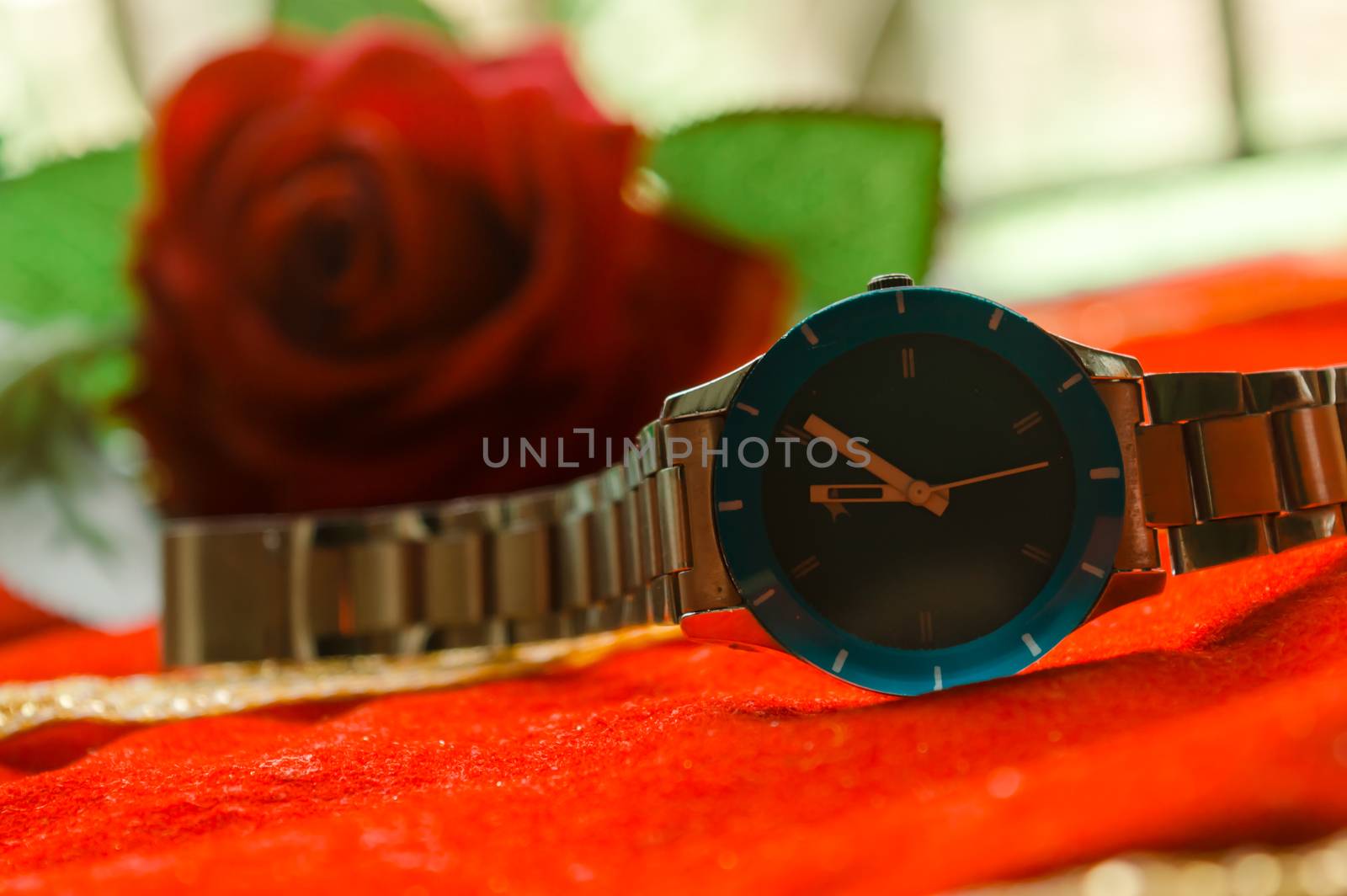 Close up woman wrist watch on red satin cloth. Soft focus beautiful red rose in background. Side view. Red fashion luxury elegance background. Still life.