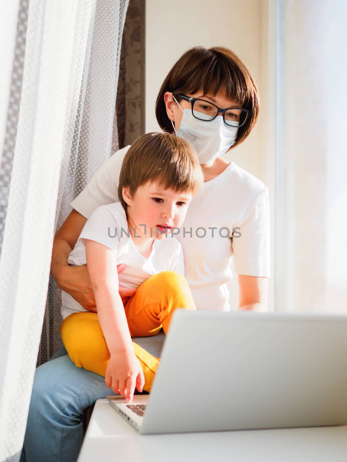 Woman in medical mask works remotely from home. She sits on window sill with laptop and cute toddler boy on her knees. Lockdown quarantine because of coronavirus COVID19. Self isolation at home.