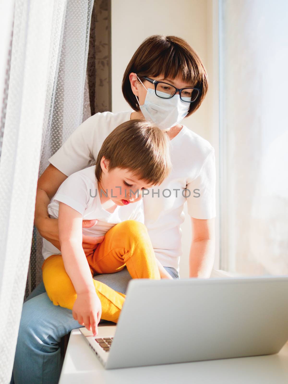 Woman in medical mask remote works from home. She sits on window sill with laptop and cute toddler boy on her knees. Lockdown quarantine because of coronavirus COVID19. Self isolation at home.