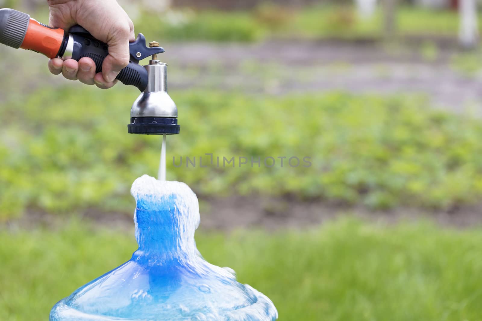 The gardener holds an irrigation sprinkler and collects clean water in a blue bottle against a blurred bright green grass.