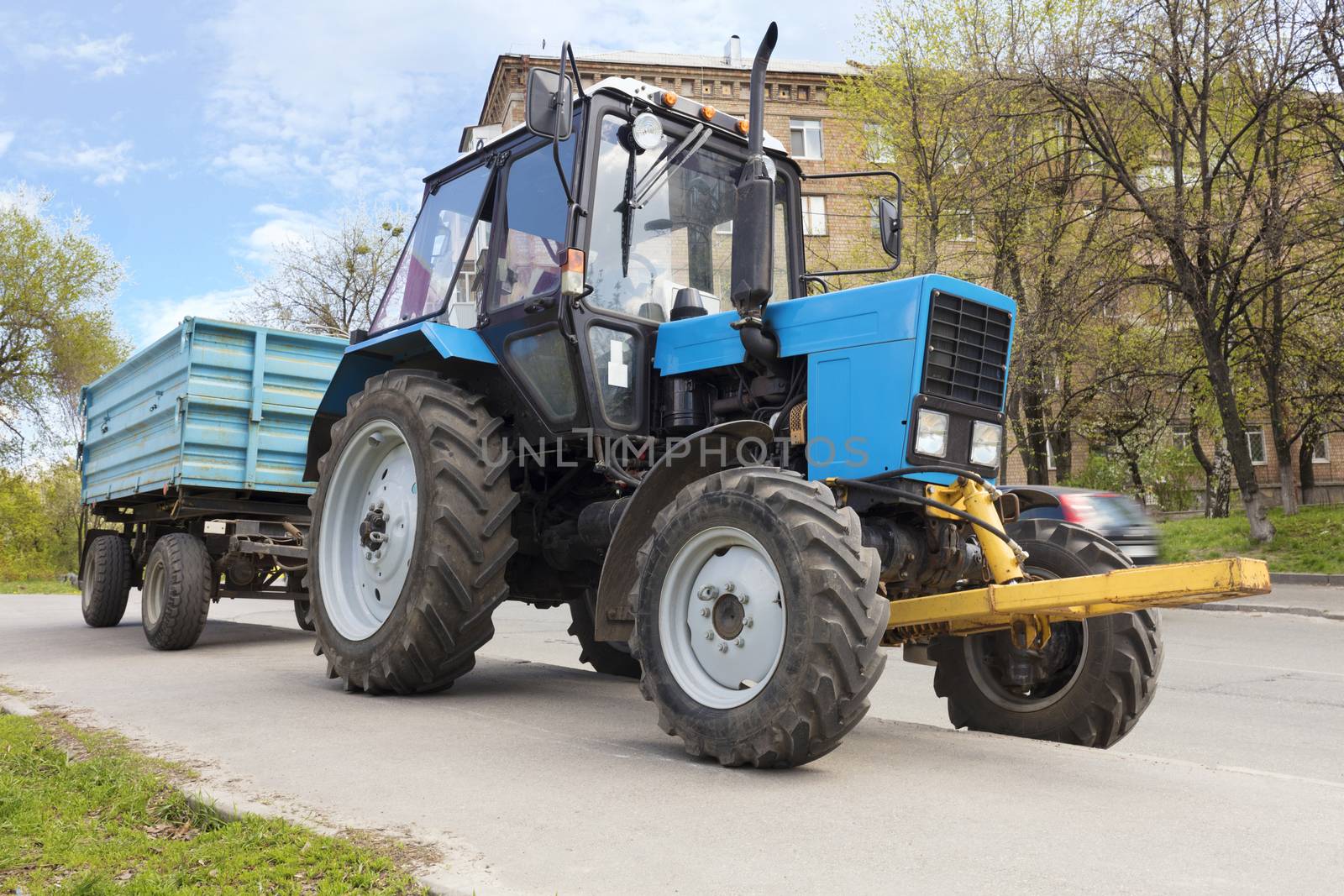 An old small blue tractor with a trailer stands on the edge of a city street road