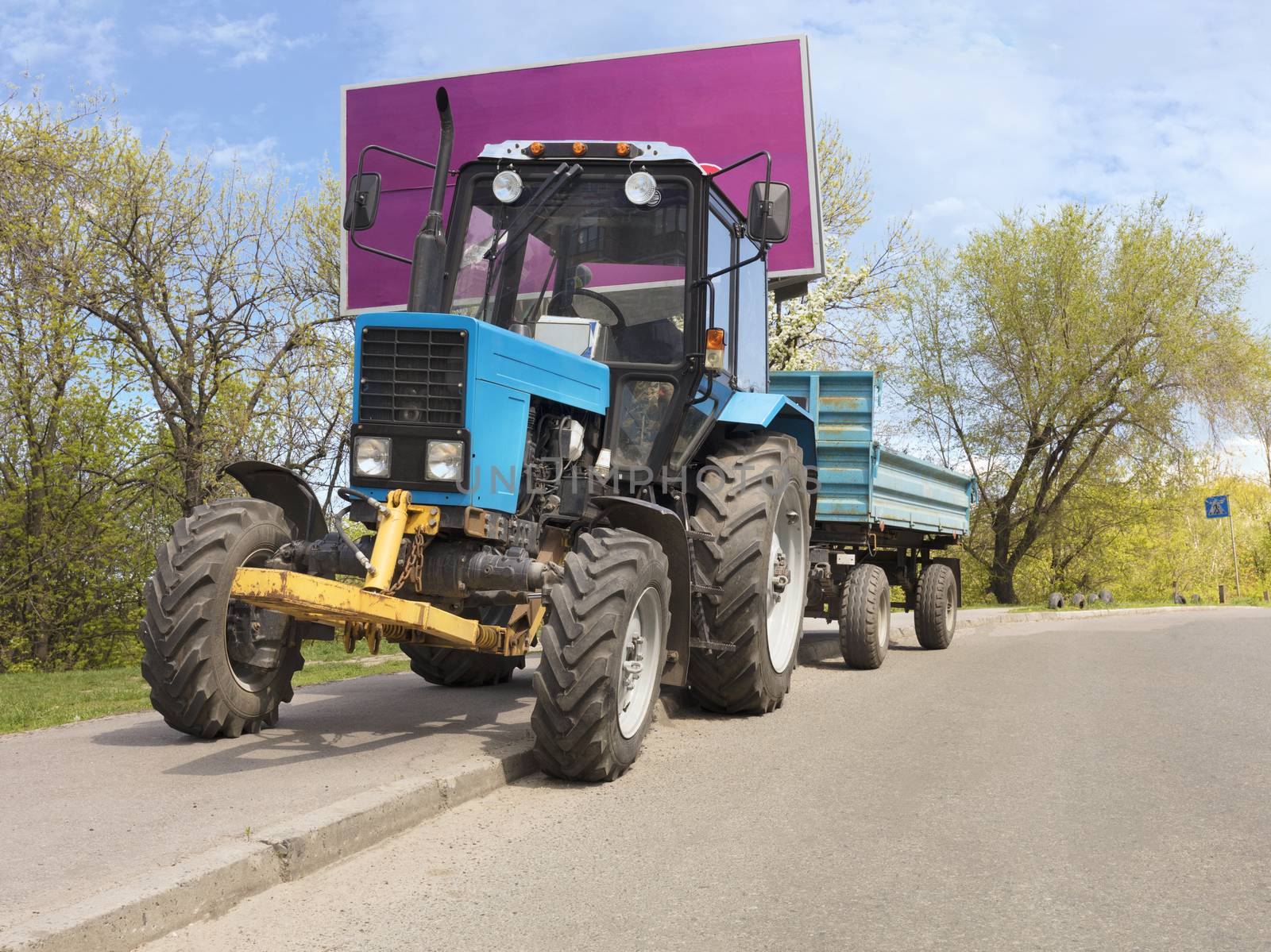 An old small blue tractor with a trailer stands on the side of the road against the background of a green spring park near a large billboard. Place for text.
