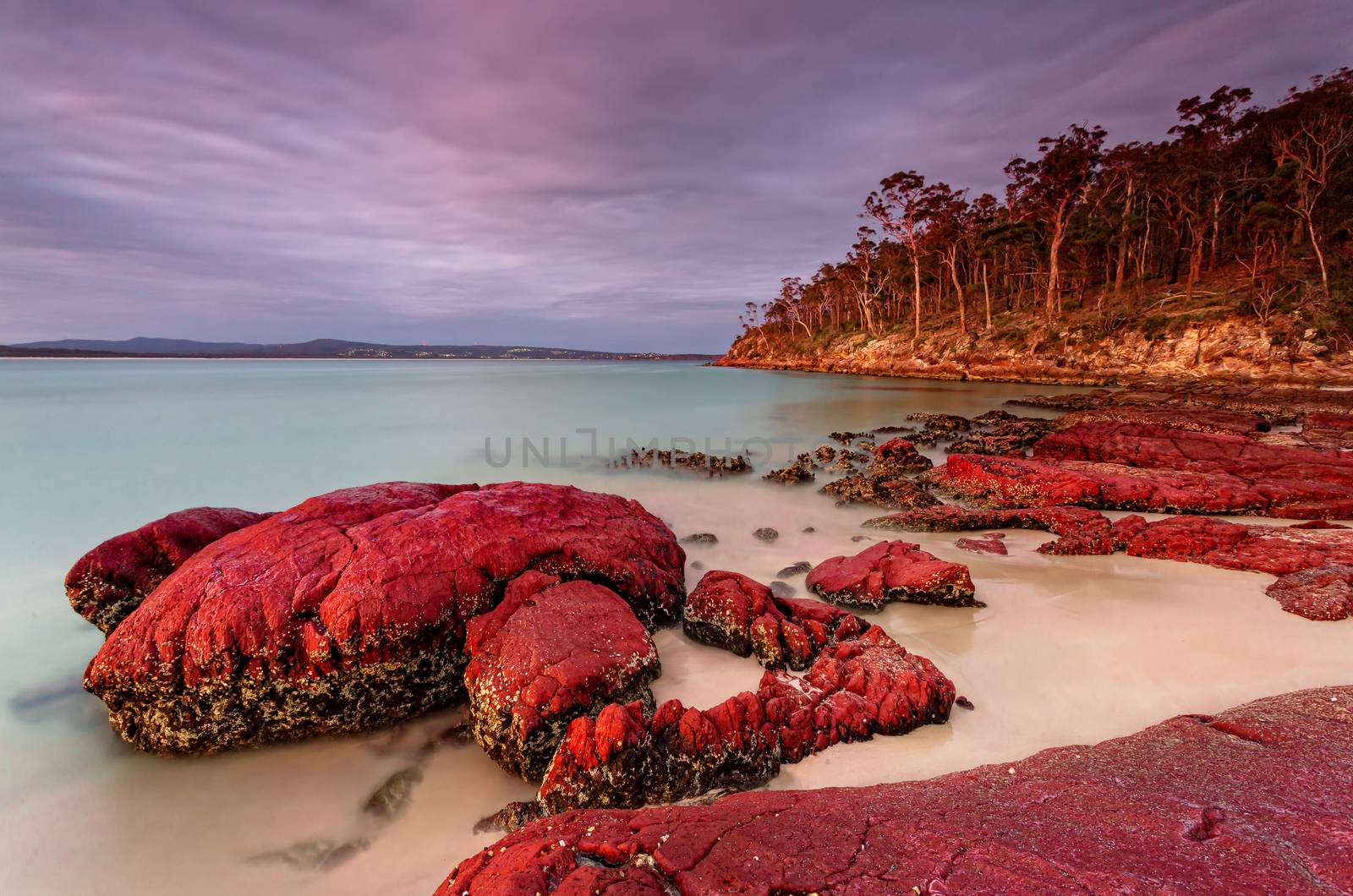 Sunset casting warm light onto the rich red rocks scattered along the beach contrasting against the blue of the waters