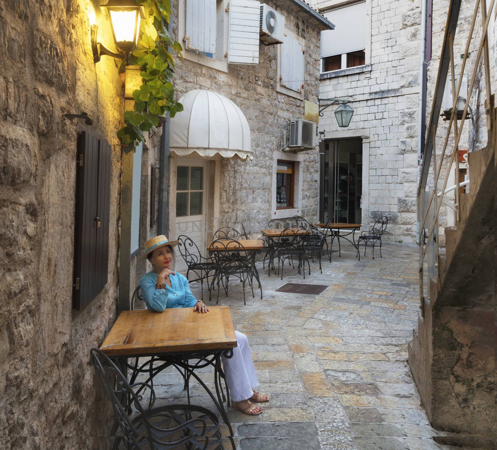 A young woman in a bright turquoise blouse and a straw hat sits at a table of a street cafe in the old town of Budva, Montenegro