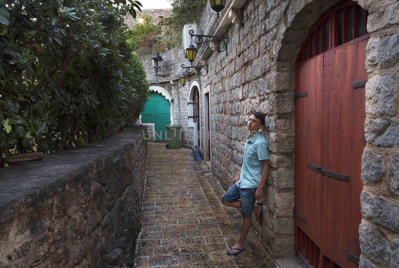 The young man stands near the wooden gate in the narrow street of the old town of Budva, Montenegro. by Sergii