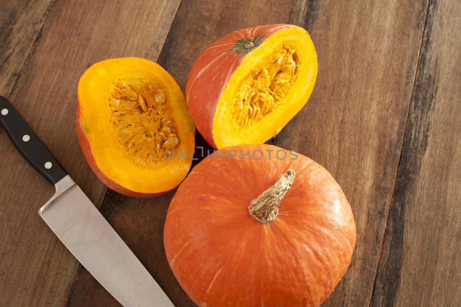 Small whole and halved colorful orange fall pumpkin or squash with a large kitchen knife on a wooden kitchen table, high angle view