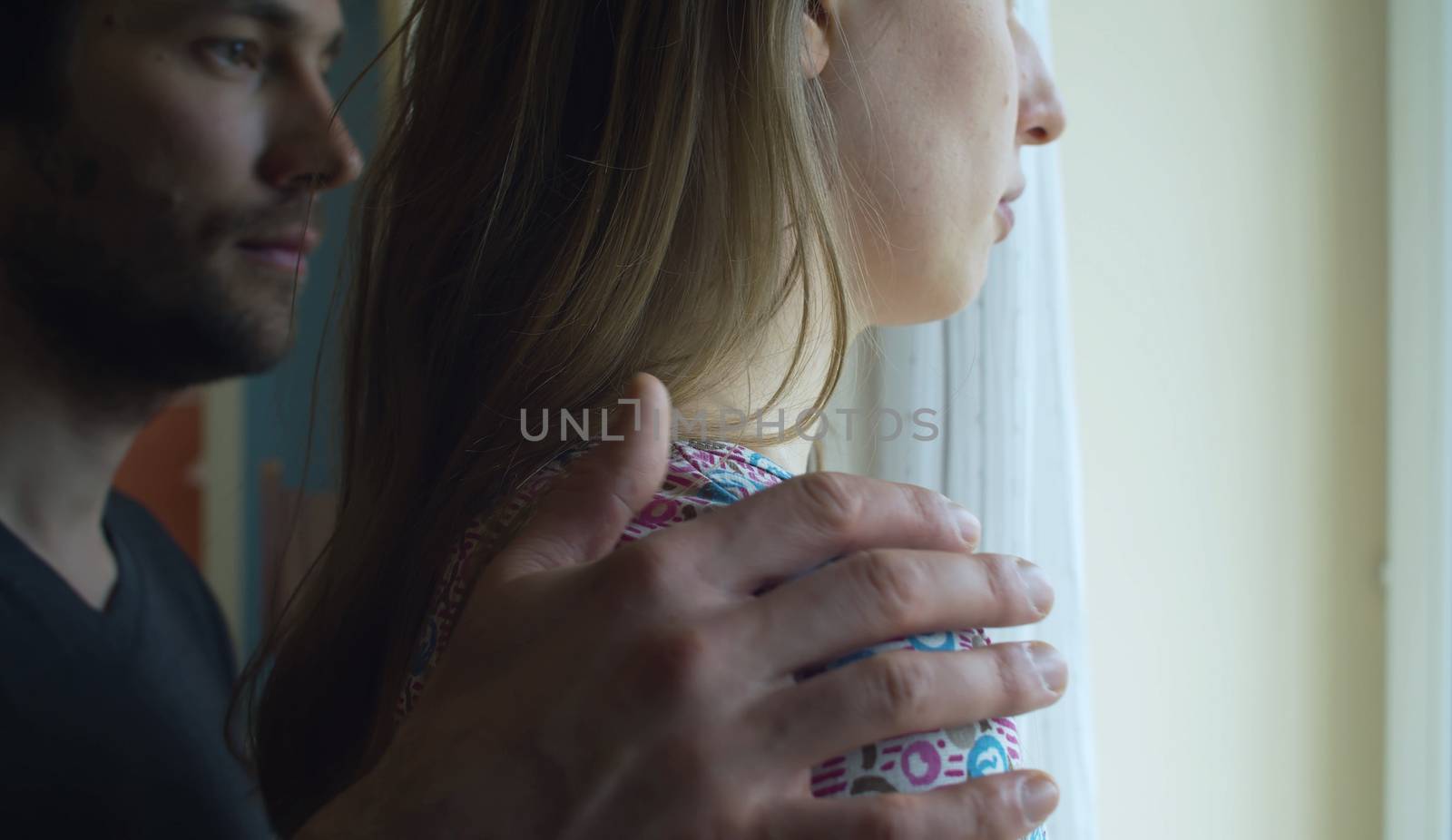 Close up portrait of a sad woman in profile near the window. Her husband puts hand on her shoulder in search of reconciliation.