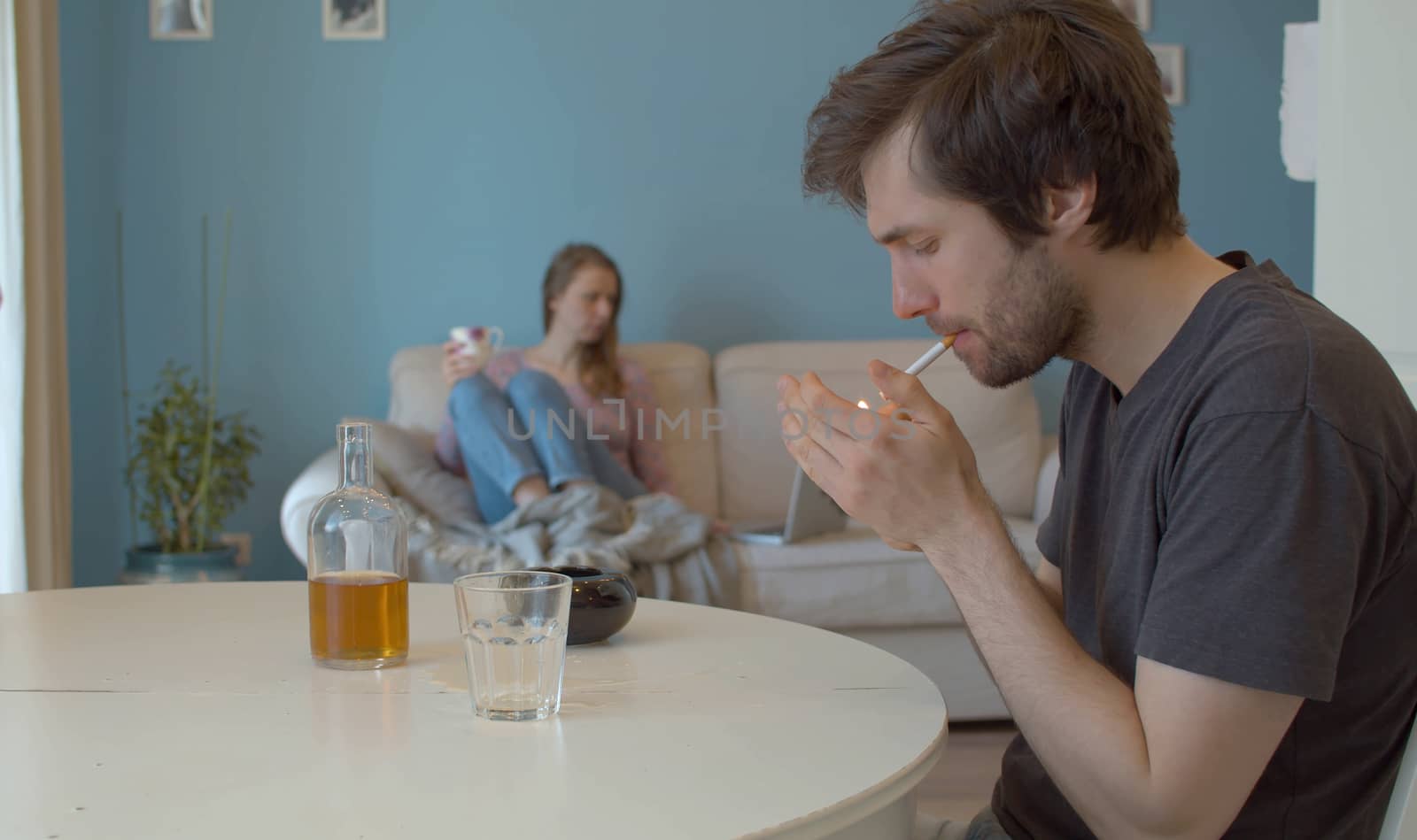 A drunk, sad man lighting a cigarette. In the background, in defocus, the wife sits on the couch and looks at the computer.